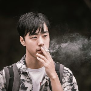 An adult male exhaling smoke while smoking outdoors in casual clothing.